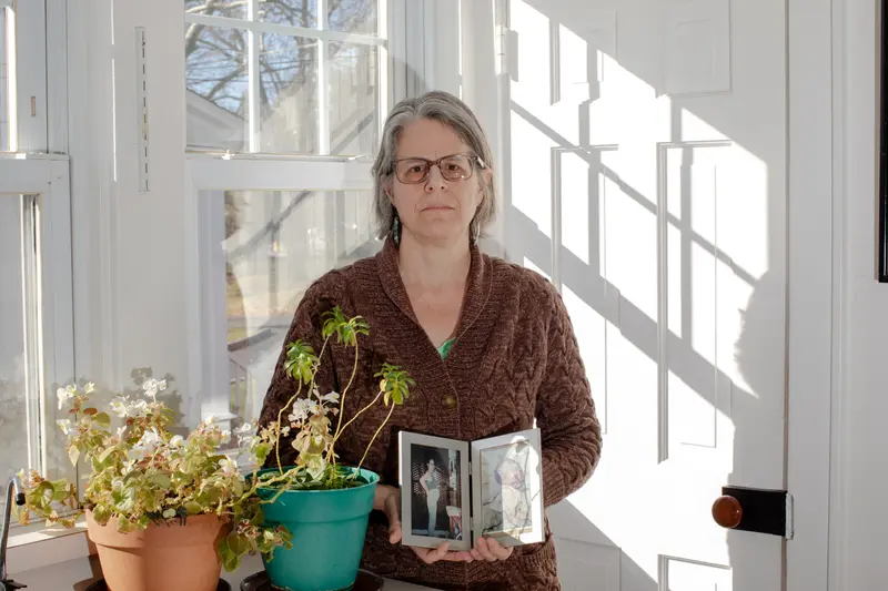 A white woman in a brown cardigan holds a frame with two photos.